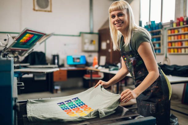 Happy print shop worker putting screen printed t-shirt in drying machine Portrait of a smiling printing workshop female worker putting silkscreen printed textile product into drying machine at facility. Happy female graphic technology expert finishing printing and drying t shirt transfer stock pictures, royalty-free photos & images