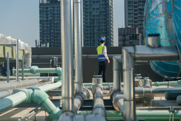 istock Focused engineer in hard hat and reflective vest standing amidst rooftop mechanical structures, consulting blueprints. 2151804378