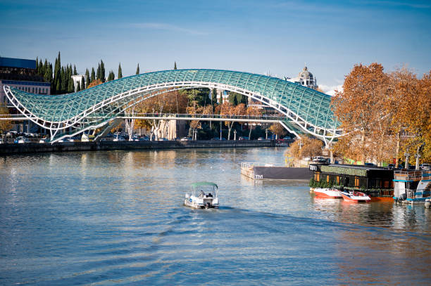 istock Peace Bridge, a modern architectural marvel, stretching across the Kura River in Tbilisi. The autumnal trees along the riverside add a seasonal charm, while boats navigate the tranquil waters, 2151835295