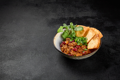 Chilli con carne with beans and corn tortilla on black background. Traditional mexican food - chilli with minced meat and spices in bowl on dark backdrop. Chili con carne in minimal style