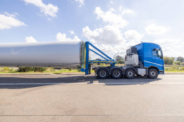 Captured on a sunny day with fluffy clouds above, this image shows a vivid blue semi-truck transporting a massive wind turbine blade. The blade is mounted on a specialized trailer designed to handle its extraordinary length and weight, emphasizing the essential role of logistics in the renewable energy sector.