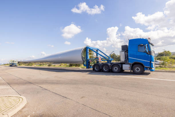 A blue semi-truck with a specialized trailer transporting an extraordinarily long wind turbine blade along a highway. The photograph showcases the vast scale of the turbine blade against the backdrop of a clear blue sky, highlighting the critical and innovative logistical approaches required to support the renewable energy industry.