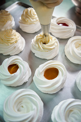 Top view preparation of delicious appetizing mini Pavlova dessert, confectioner adding whipped cream-cheese on the top of merengue nests with fruit mousse inside