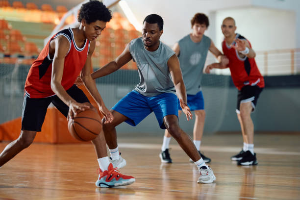 Black defense player trying to block his opponent during basketball match. Male athletes playing basketball match in school gymnasium. youth basketball indoor stock pictures, royalty-free photos & images
