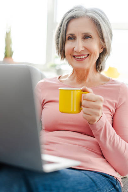 istock Vertical view of happy senior woman using laptop computer, holding cup, sitting on sofa 2151852967