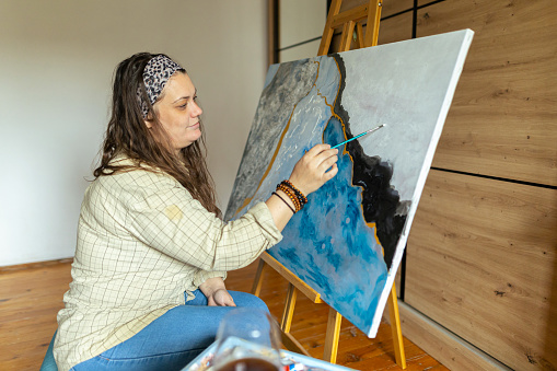 An confidence, artist paints her picture in her studio. He wears a light plaid shirt. Thoughtful, concentrated and happy, she finishes her work. Real life photo, real woman expressing herself in art . Copy space