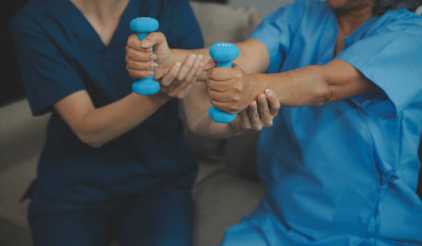 istock Old woman training with physiotherapist using dumbbells at home. Therapist assisting senior woman with exercises in nursing home. Elderly patient using dumbbells with outstretched arms. 2151865405