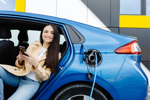 istock Business lady sitting inside charging electric car and using modern smartphone. Technology and transport concept. 2151867975
