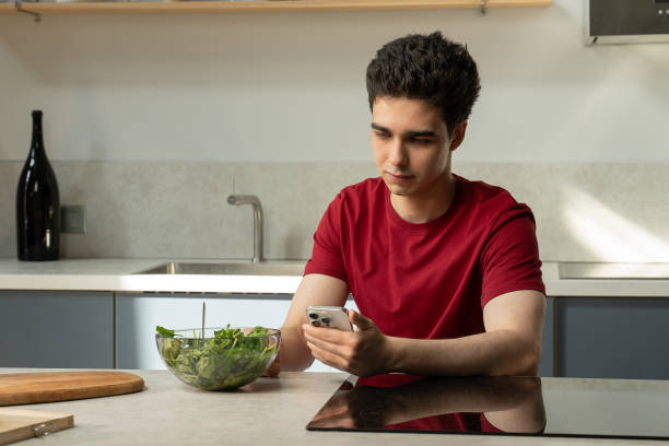 istock Young Man Sitting at Kitchen Table Looking at Cell Phone 2151877760