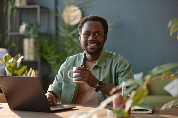 Smiling Black Man Sitting at Workplace in Office Decorated with Plants