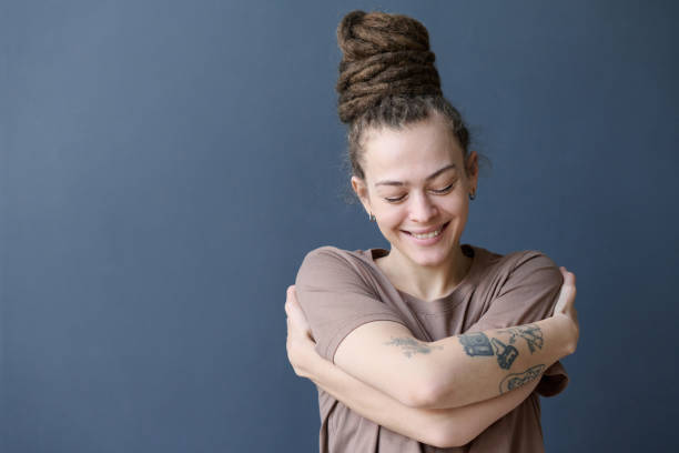 Smiling Woman with Dreadlocks in Bun Embracing Herself Standing by Grey