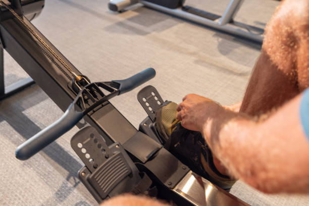 A close-up view of a man adjusting his footwear on a rowing machine, showcasing the details of his pre-workout setup. The focus is on his hands securing the straps over his shoes, emphasizing the importance of proper equipment use for effective training. The rowing machine handle, footrest, and sliding seat are visible, highlighting the essential components of the machine that enable a full-body workout. The environment is a well-equipped gym with a focus on cleanliness and modernity, seen in the minimalist design and neutral color palette.
