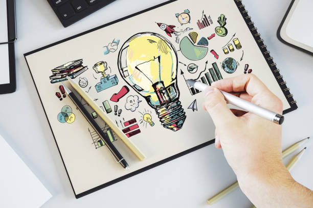 istock A person's hand drawing a creative concept with a glowing light bulb on paper, symbols and graphs, on a white desk 2151895974