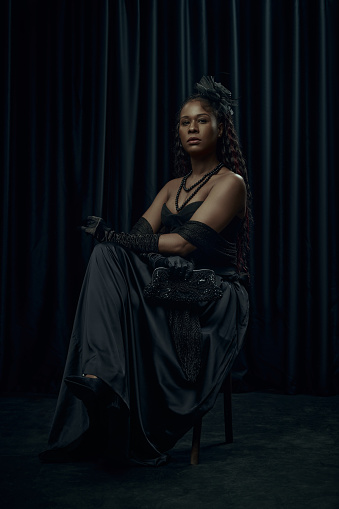 African-American woman, dressed old-fashion dress, looks as princess, sits confidently on chair against dark curtain backdrop. Concept of comparison of eras, fashion and style, modernity and history.