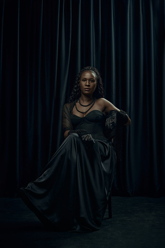 African-American woman with long, braided hair sits confidently on chair, draped in old-fashion flowing black dress against dark curtain backdrop. Concept of comparison of eras, modernity and history.