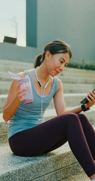 istock Young Asian female athlete runner drinking protein shake and using smartphone after exercise workout in urban park. Diet and healthy food workout exercise. Vertical Screen. 2151903571