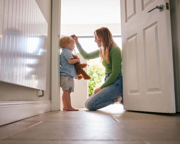 mother measuring height of young son holding teddy bear at home against kitchen door frame - groeimeter fotos stockfoto's en -beelden