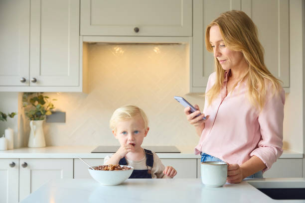 istock Mother At Home Looking At Mobile Phone As Young Son Eats Breakfast In Kitchen 2151909624