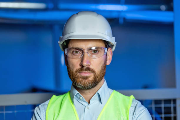 istock Handsome skilled bearded engineer in hardhat,goggles and vest looking at camera on blue background 2151919130