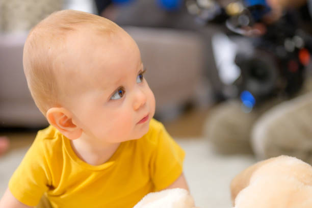 A curious baby in a yellow top sits at home with a toy, gazing upward, while a parent in casual attire sits nearby, blurred in the background.