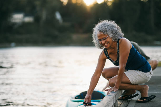 Cheerful senior woman preparing to kayak on river at sunset A vibrant senior woman of Pacific Islander descent smiles while crouching down on a river dock to lower a kayak into the water, on a beautiful summer evening in Portland, Oregon. active seniors stock pictures, royalty-free photos & images