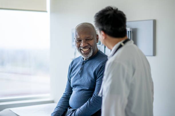 Senior Medical Exam A senior gentleman of African decent sits up on an exam table during a routine medical exam. His doctor is standing to the side of him in a white lab coat as they discuss his health. patient stock pictures, royalty-free photos & images