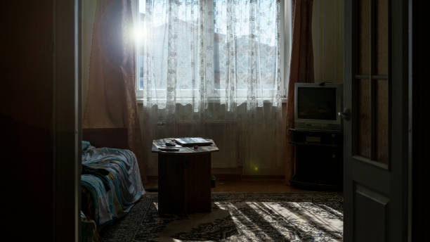 Sunlight filters through lace curtains in cozy living room casting patterns on carpet and furniture. Old television and notebook on table capture quiet nostalgic atmosphere.