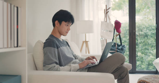 istock Young Asian Man Using Laptop in Bright Living Room. 2151985435