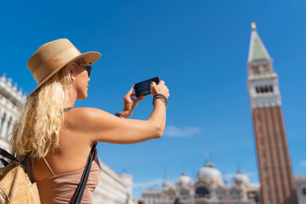 istock Female traveler with hat and backpack using smartphone and standing on San Marco square with tower and basilica on the background in Venice, Italy 2151995290