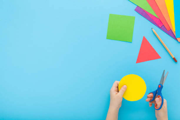 istock Little child hands holding scissors and cutting colorful geometric shapes from application paper on pastel blue table background. Making different forms. Point of view shot. Closeup. Top down view. 2152003452