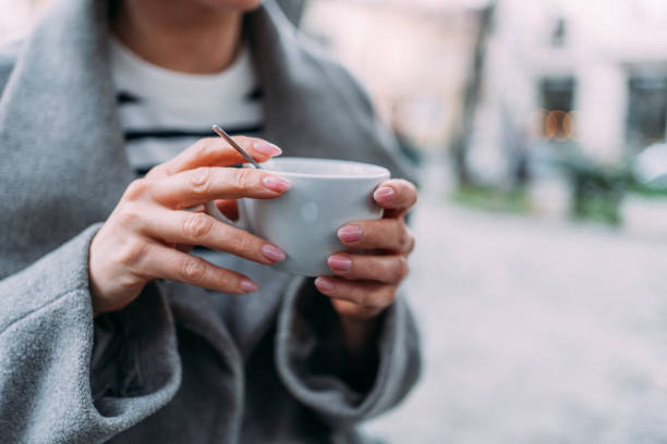 Senior woman drinking coffee in autumn cafe outdoors. Retired person eating coffee cream with caramel and enjoying morning. Cozy table with fallen leaves. Close up of female hands with cup and spoon.
