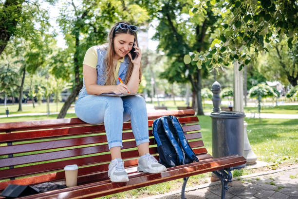 istock Young woman working outside office in public park while sitting on park bench and talking on mobile phone 2152024307