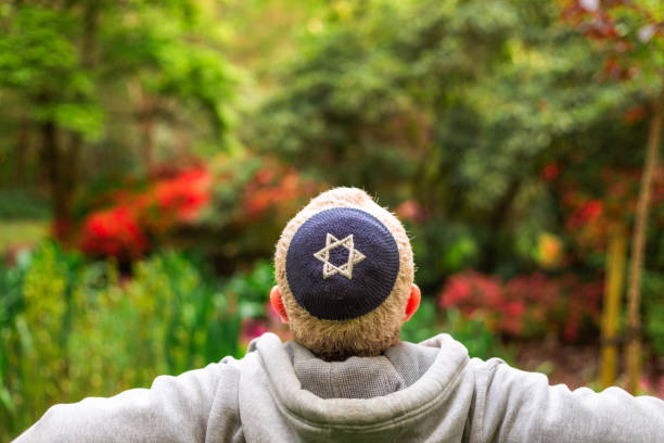 Color image depicting the rear view of a Jewish man wearing a blue skull cap (or Kippah) embroidered with the Star of David while sitting alone on a bench in the park. Focus on the man with the lush foliage of the park defocused beyond.