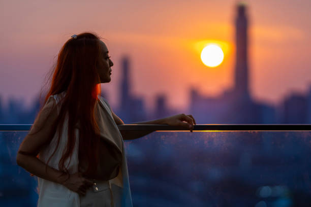 istock Woman looking and enjoying the sunset view from balcony with the sun setting behind skyscraper in busy urban downtown with loneliness for solitude, loneliness and dreaming of freedom lifestyle 2152045255