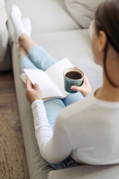 istock Young woman sit on sofa, hold coffee cup in hand, read paper book 2152046811