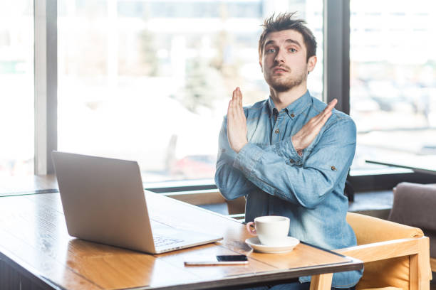 istock Serious handsome man freelancer working on laptop, showing x sign, no way gesture, looking at camera 2152049009