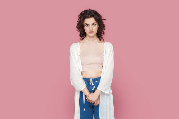 Portrait of serious concentrated woman with curly hair wearing casual style outfit standing looking at camera with attentive expression. Indoor studio shot isolated on pink background.