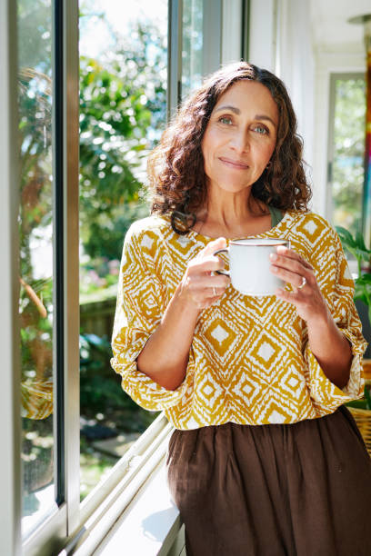 istock Mature woman smiling while drinking coffee by an open window at home 2152059870