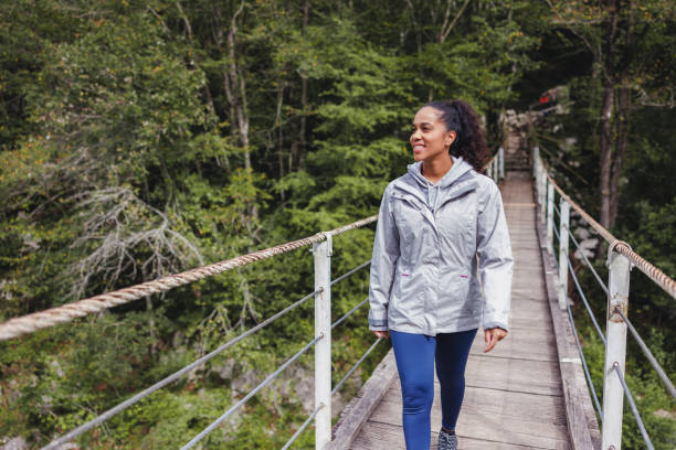 A young adult woman walks across a rustic suspension bridge in a lush green forest, smiling and enjoying the outdoors.