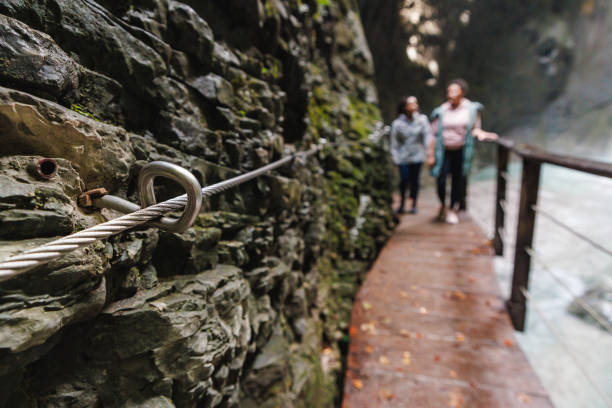 Two women walking along a wooden walkway beside a rocky cliff, exploring the natural beauty of Kozjak Waterfall in Kobarid.