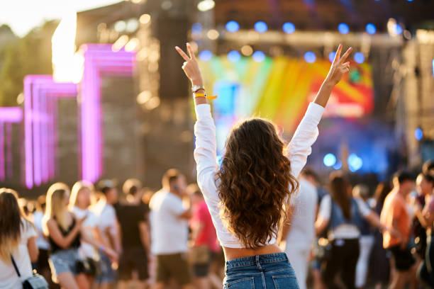 Raised hands with peace sign, enjoying live concert. Back view of young woman at summer beach music festival. Crowd, stage lights, sunset party vibes. Casual fashion, outdoor event, fun atmosphere. vista trasera de una mujer joven en el festival de música de la playa de verano. manos levantadas con el signo de la paz, disfrutando de un concierto en vivo. multitud, luces del escenario, vibraciones de fiesta al atardecer. moda casual, evento al aire l