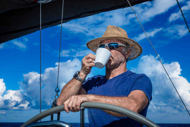 A man in a straw hat drinks coffee while driving a sailboat.