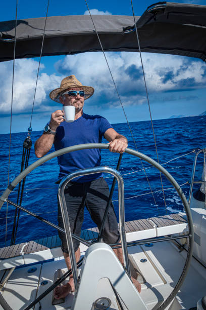 A man in a straw hat drinks coffee while driving a sailboat.