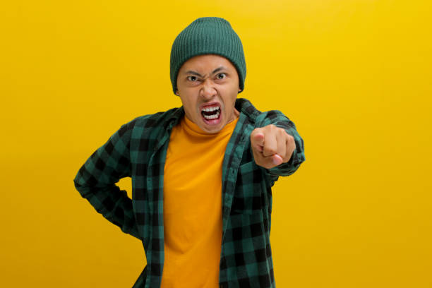 Angry young Asian man is yelling, pointing a finger, blaming all failures on someone Angry young Asian man in a beanie hat and casual shirt is yelling, pointing a finger, and expressing his frustration, blaming all failures on someone. Isolated on yellow background. asian suit man cross hand stock pictures, royalty-free photos & images