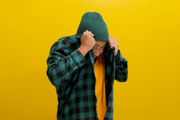 The amusingly frightened young Asian man hides his face with his shirt, expressing a mix of fear, anxiety, and shyness, while standing against a yellow background