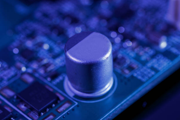Close-up view of an electronic component on a circuit board. The component appears to be a silver cylindrical capacitor, surrounded by other circuitry elements. The boards surface is green