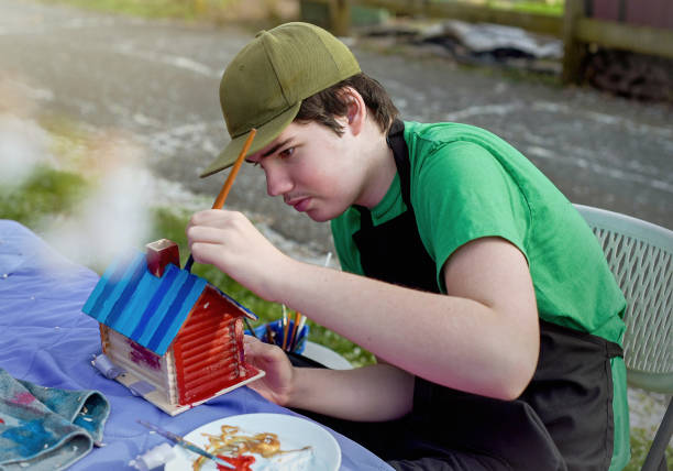 18-year-old boy from Madison, New Jersey, with autism meticulously paints a colorful birdhouse in a backyard setting. The focus and precision in his brushwork illustrate deep concentration and a creative outlet that aids in his expression and enjoyment.
