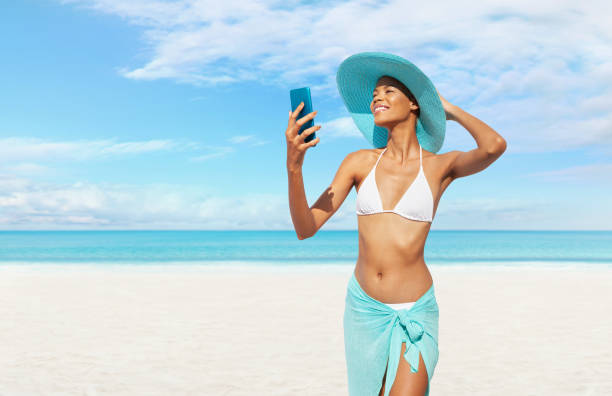 istock Happy young woman at the beach side using mobile phone, wearing a turquoise sun hat and bikini, portrait of African latin American woman in sunny summer day with blue sky, concept of a summer holiday 2152135292