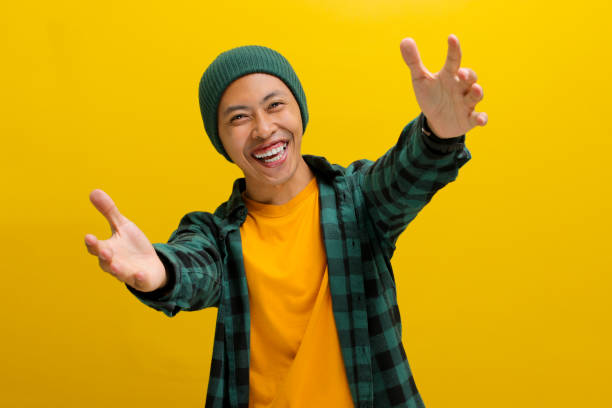 Overjoyed Asian man stretches his arms wide open towards the camera Overjoyed Asian man, dressed in a beanie hat and casual shirt, stretches his arms wide open towards the camera, offering a hug gesture, inviting an embrace while standing against a yellow background male is standing and shows outstretched hand with open palm stock pictures, royalty-free photos & images