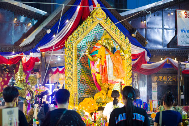 Golden Ganesha statue at night at Ganesha shrine and temple in Bangkok Huai Khwang at Ratchadaphisek Road. People are standing in front of golden statue and are praying. One woman is standing close to statue and is placing incense sticks. rear view of people standing in rows